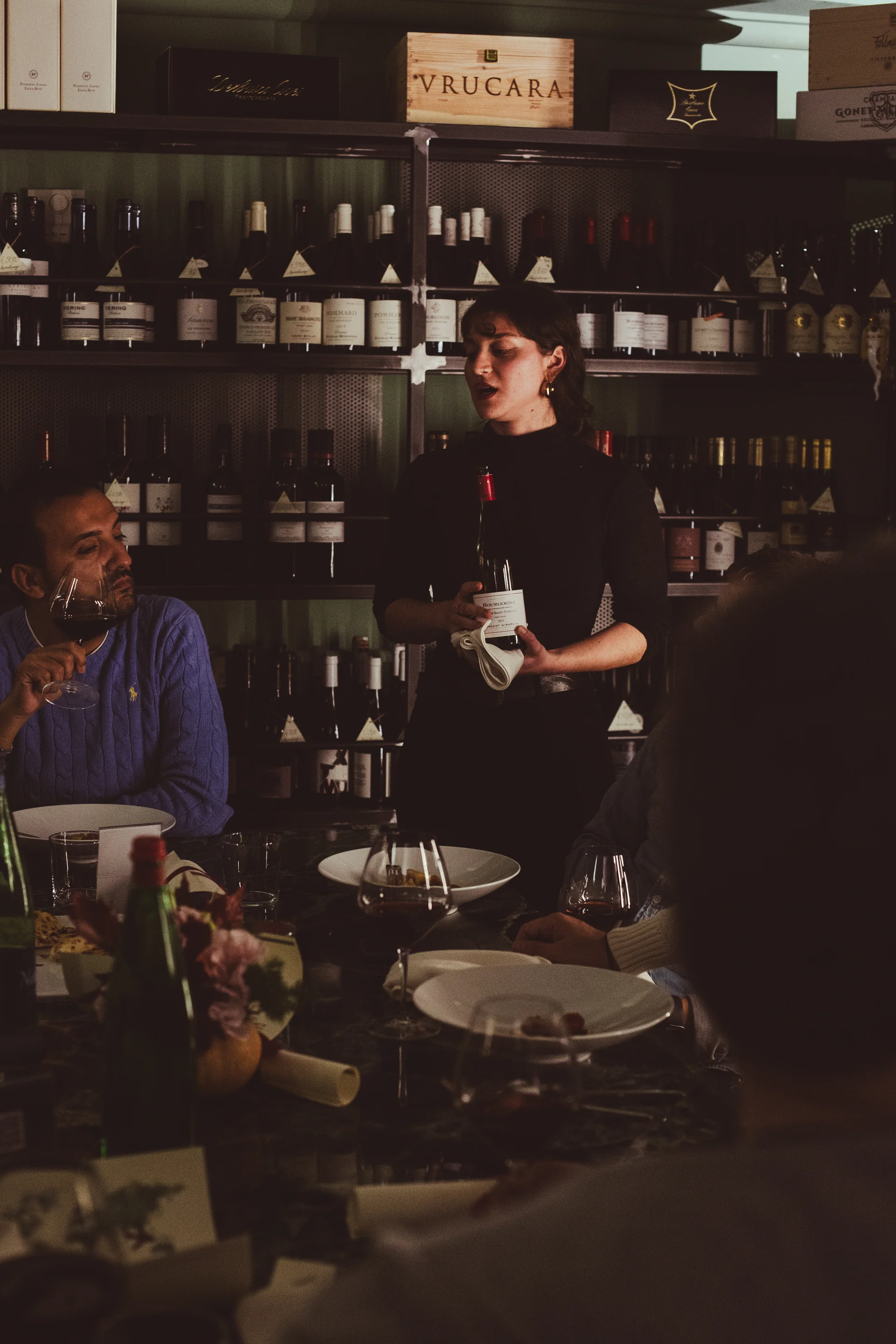 Sommelier che presenta una bottiglia di vino durante una degustazione guidata al bancone, con ospiti seduti che assaggiano dai calici